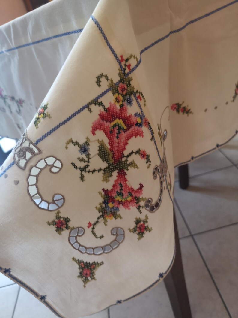Puede incluir: Un mantel blanco con un borde azul y dise&ntilde;os florales bordados intrincados en tonos de rosa, rojo, verde y azul. El bordado presenta un patr&oacute;n repetitivo de flores y hojas.