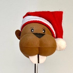 May include: A brown beaver wearing a red and white Santa hat with a white pom-pom. The beaver has a white tooth and is attached to a silver metal rod.