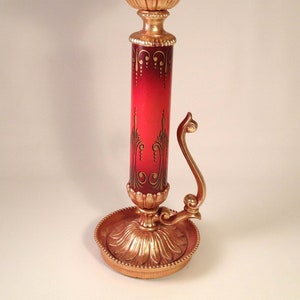 May include: A gold candle holder with a red and gold base and a decorative design. The candle holder has a curved arm and a round base.