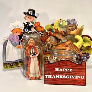 May include: A Thanksgiving themed papercraft scene with a glass jar filled with a pilgrim woman figurine, a pheasant figurine, and a sign that reads "Happy Thanksgiving".