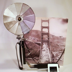 May include: A vintage camera flash with a silver reflector and a black handle. The flash is attached to a tripod with a black strap. The background is a black and white photo of the Golden Gate Bridge.