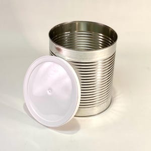 May include: A cylindrical metal storage container with horizontal ridges along the exterior walls and a smooth, circular white plastic lid resting upright against the base of the open-topped canister, which features a metallic silver finish.