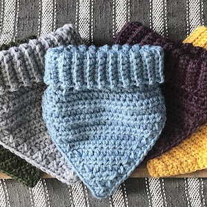 May include: Four crocheted dog bandanas in different colors: green, gray, blue, and yellow. The bandanas are folded and arranged on a wooden surface.