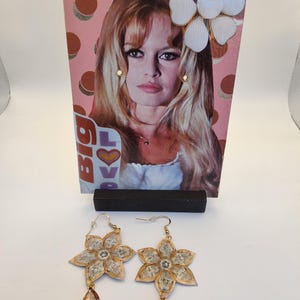 May include: A pair of flower-shaped earrings with gold-colored edges and clear centers, each with a teardrop-shaped, gold-colored pendant. A vintage magazine clipping featuring a woman with blonde hair is in the background.