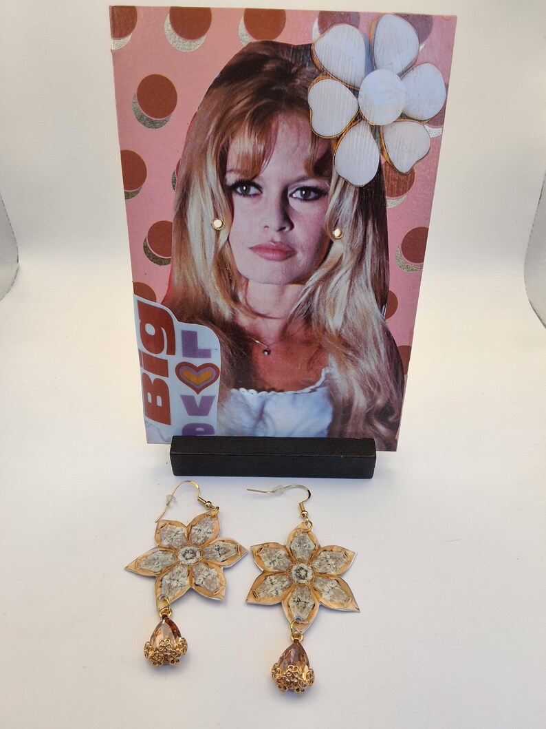 May include: A pair of flower-shaped earrings with gold-colored edges and clear centers, each with a teardrop-shaped, gold-colored pendant. A vintage magazine clipping featuring a woman with blonde hair is in the background.