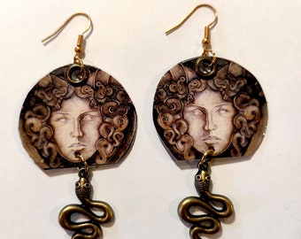 May include: A pair of gold earrings with a round, brown and white image of Medusa's face with snakes for hair. Each earring has a dangling bronze snake charm.