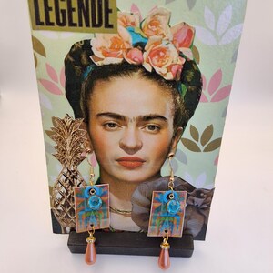 Peut inclure: Boucles d'oreilles dorées avec des accents de pierres bleues et des pampilles en forme de goutte d'eau pêche. Les boucles d'oreilles sont présentées devant un présentoir décoratif avec un portrait de Frida Kahlo et le mot "LEGENDE".