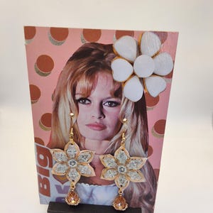 May include: Floral earrings with gold-tone metal and a teardrop-shaped amber-colored gem. The earrings are flower-shaped with a light-colored center. A matching flower-shaped brooch is also visible. The background features a vintage magazine cover.