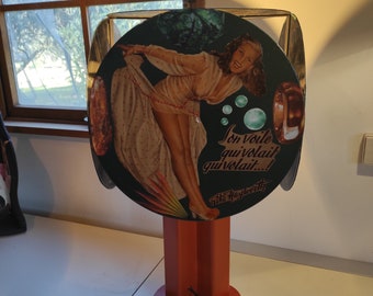 Puede incluir: Lámpara de estilo vintage con una ilustración de chica pin-up en una pantalla circular. La pantalla tiene un fondo oscuro con texto y una imagen de una mujer con un vestido. La lámpara tiene una base geométrica roja y un cable de alimentación negro.