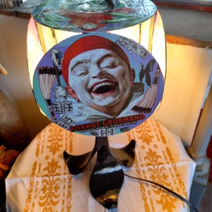 May include: A black metal lamp base with a round, colourful lampshade. The lampshade features a vintage illustration of a laughing clown with the text "PIERROT GOURMAND CHOCOLATIER CONFISEUR".