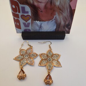 May include: A pair of gold-colored flower earrings with a teardrop-shaped charm. The earrings have a floral design with a textured, light-colored center and a gold-colored border. The teardrop charm is a light orange color.