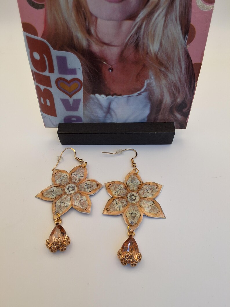 May include: A pair of gold-colored flower earrings with a teardrop-shaped charm. The earrings have a floral design with a textured, light-colored center and a gold-colored border. The teardrop charm is a light orange color.