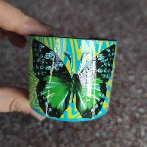 Pulsera de cartón de una mariposa con purpurina sobre un fondo de papel verde y azul vintage en un collage de papel recortado "MARIPOSA"