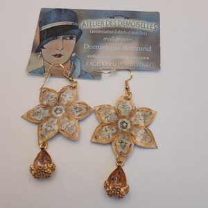 May include: A pair of handmade flower earrings. The earrings feature a gold-colored hook, a flower-shaped design with a beige and white pattern, and a teardrop-shaped charm with gold-colored beads. The earrings are displayed on a white surface.
