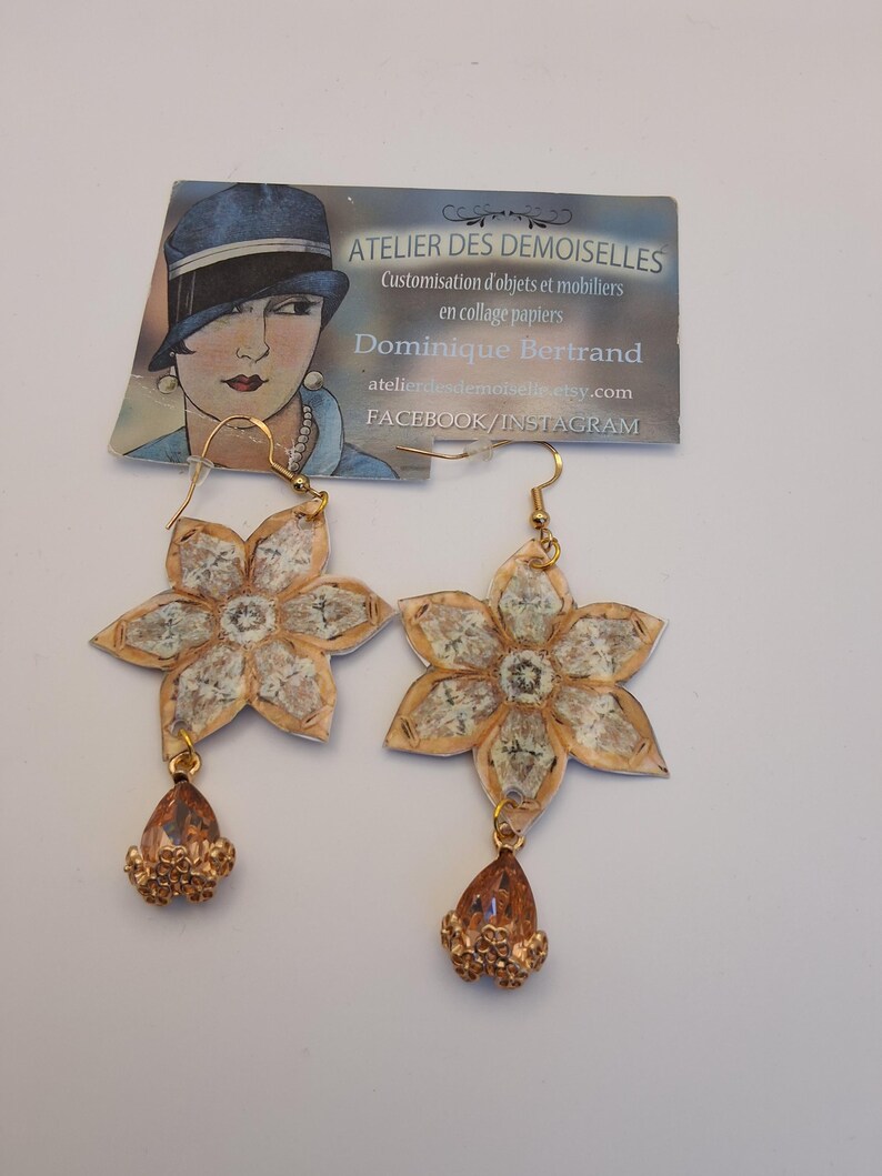 May include: A pair of handmade flower earrings. The earrings feature a gold-colored hook, a flower-shaped design with a beige and white pattern, and a teardrop-shaped charm with gold-colored beads. The earrings are displayed on a white surface.