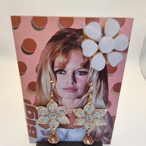 May include: Floral dangle earrings with a gold-tone finish and a peach-colored teardrop gem. The earrings are flower-shaped with a light-colored center. A large white flower with gold trim is attached to a pink background with a woman's face.