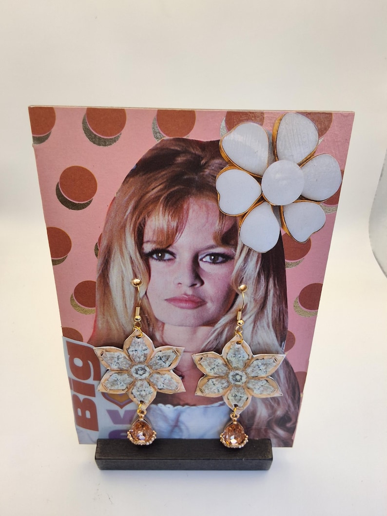 May include: Floral dangle earrings with a gold-tone finish and a peach-colored teardrop gem. The earrings are flower-shaped with a light-colored center. A large white flower with gold trim is attached to a pink background with a woman's face.