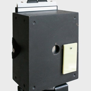 Könnte beinhalten: Eine rechteckige schwarze Boxkamera mit einer zentralen kreisförmigen Objektivöffnung, einer hellgelben rechteckigen Abdeckplatte, die mit einer Schraube befestigt ist, und einem weißen, oben montierten Filmhalter. Das Gerät hat einen schwarzen Seitenriemen.
