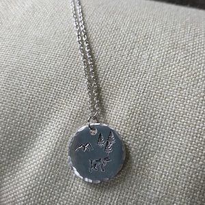 Könnte beinhalten: Eine silberfarbene Halskette mit einem runden Anhänger mit gehämmertem Rand. Der Anhänger ist mit einem kleinen Gebirge, zwei Nadelbäumen und der Silhouette eines Hundes graviert. Er hängt an einer zarten, dünnen silberfarbenen Metallkette.