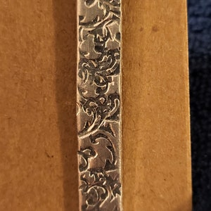 Puede incluir: Un colgante de plata con un diseño floral detallado. El colgante es rectangular y tiene un acabado liso y pulido.