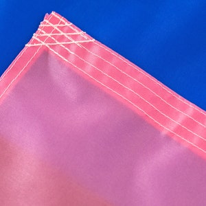 May include: Close-up of a pink fabric with white stitching. The fabric is part of a larger flag with a blue section visible in the background.