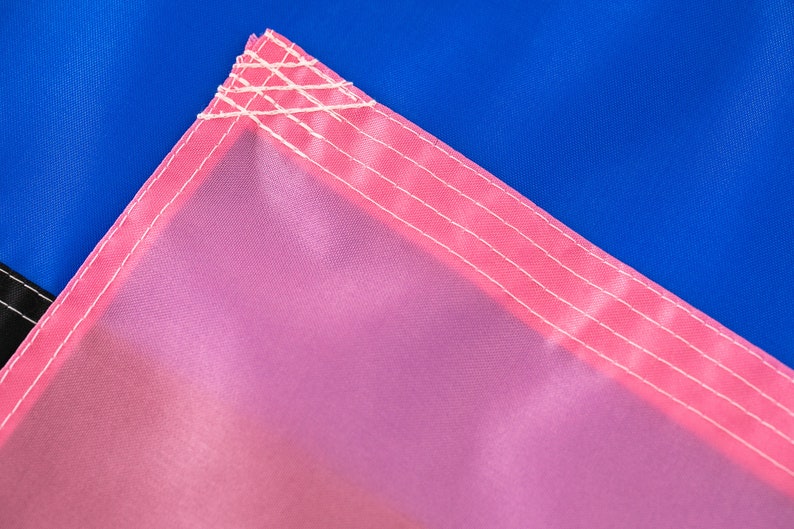 May include: Close-up of a pink fabric with white stitching. The fabric is part of a larger flag with a blue section visible in the background.