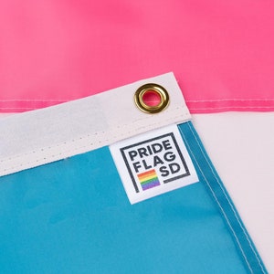 May include: Close-up of a blue, white, and pink flag with a sewn-on label that reads "PRIDE FLAG SD" with a rainbow stripe below the text.