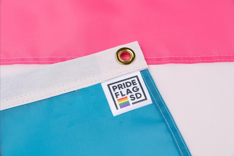 May include: Close-up of a blue, white, and pink flag with a sewn-on label that reads "PRIDE FLAG SD" with a rainbow stripe below the text.