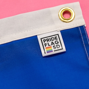 May include: Close-up of a blue, white, and pink flag with a white label that says "PRIDE FLAG SD" and a rainbow stripe. The label is attached to the blue section of the flag.