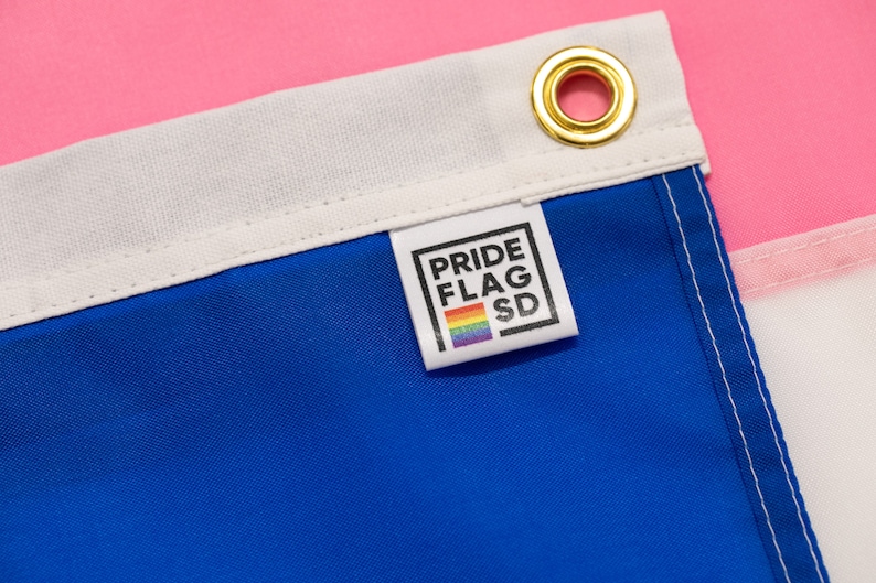May include: Close-up of a blue, white, and pink flag with a white label that says "PRIDE FLAG SD" and a rainbow stripe. The label is attached to the blue section of the flag.