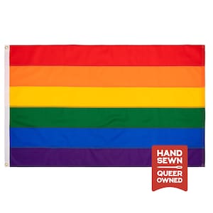 May include: A rectangular pride flag featuring six horizontal stripes in red, orange, yellow, green, blue, and purple. A white header strip with two metal grommets is on the left side. A red tag in the bottom right corner reads HAND SEWN QUEER OWNED.