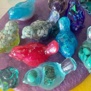 Puede incluir: Una colección de coloridas figuras de pájaros de resina en varios tonos de azul, verde, rojo y morado. Algunas aves están rellenas de piedras o purpurina. Las figuras se exhiben sobre una superficie morada.