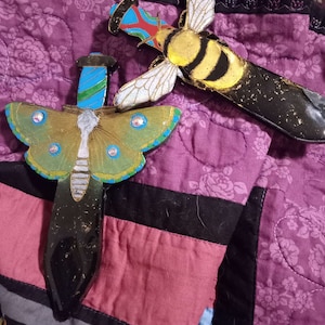May include: Two decorative, hand-painted, black dagger-shaped ornaments. One features a green and blue butterfly with iridescent wings. The other features a yellow and black bee with gold accents.
