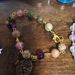 May include: A gold chain bracelet featuring a mix of green, pink and purple beads. The bracelet has a small, antique-looking clock charm attached.