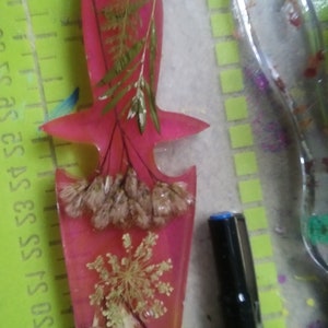 May include: A pink resin dagger pendant with dried flowers and bone fragments embedded within. The dagger has a star-shaped hilt and a pointed blade. A black marker is in the background.