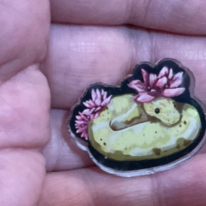 May include: A clear acrylic charm featuring a yellow and brown snake with pink flowers on its head. The snake is curled up in a circle and the flowers are in bloom.