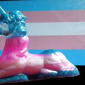 May include: A blue, pink, and white unicorn figurine with glitter, lying down on a transgender pride flag.