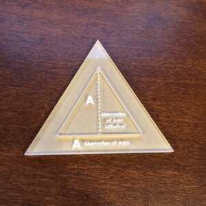 Könnte beinhalten: Eine transparente, dreieckige Quiltvorlage mit einem kleineren Dreieck im Inneren. Die Vorlage trägt den Text "A Memories of Asia Millefiori". Die Vorlage besteht aus klarem Kunststoff und liegt auf einer braunen Oberfläche.