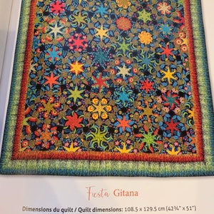 Op de afbeelding: Een kleurrijk quilt met een zwarte achtergrond en verschillende ster- en bloemenpatronen. De quilt is afgewerkt met een rode, oranje en groene rand. De quilt is gelabeld "Fiesta Gitana" en meet 108,5 cm bij 129,5 cm.