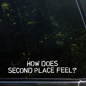 Könnte beinhalten: Weißes Vinyl-Aufkleber mit dem Text "HOW DOES SECOND PLACE FEEL?" auf einer Autoscheibe. Der Text ist in einer fetten, serifenlosen Schriftart gehalten. Der Hintergrund ist eine dunkle, reflektierende Oberfläche mit verschwommenen Bäumen.