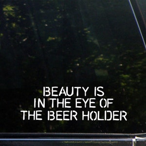 May include: A black car window with a white vinyl sticker that reads "Beauty is in the eye of the beer holder."