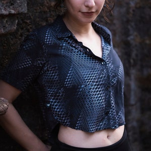 Op de afbeelding: Een vrouw met donker haar in een knot met kralen draagt een geknipt shirt met een zwart en blauw patroon en een zwarte broek.