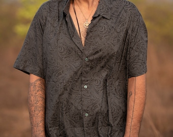 May include: A man wearing a black short-sleeved button-down shirt with a swirling pattern. He is wearing round black sunglasses and has a silver necklace with a peace sign pendant.