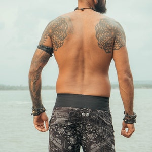 A man with tattoos on his shoulders and arms is wearing black swimming trunks with a geometric pattern. He is standing with his back to the camera, looking out at the water.