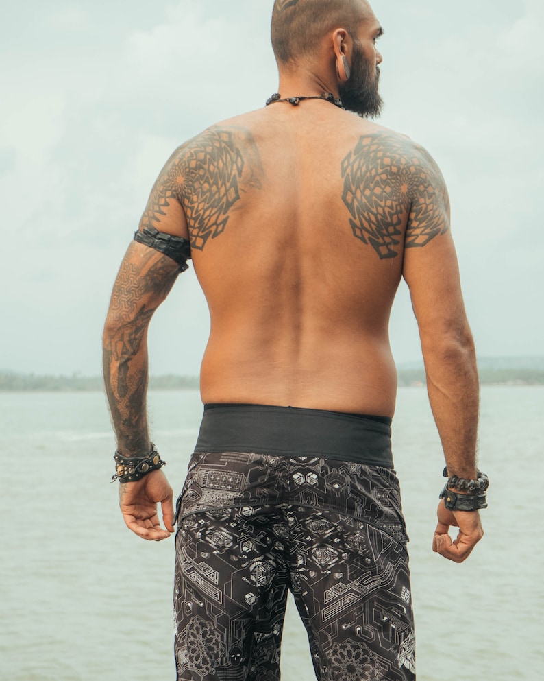 A man with tattoos on his shoulders and arms is wearing black swimming trunks with a geometric pattern. He is standing with his back to the camera, looking out at the water.