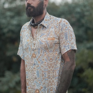 Puede incluir: Camisa de manga corta con fondo crema y estampado repetitivo en azul y naranja. La camisa tiene un bolsillo en el pecho. La persona lleva un collar de cuentas negro y una pulsera negra. La camisa está abotonada.