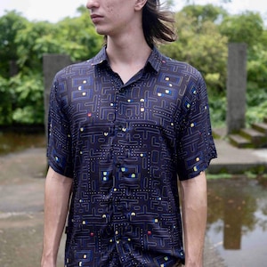 Puede incluir: Camisa azul marino de manga corta con un diseño de laberinto de Pac-Man en amarillo, azul y rojo. La camisa tiene un cuello clásico y cierre de botones. Fabricada en tejido ligero.