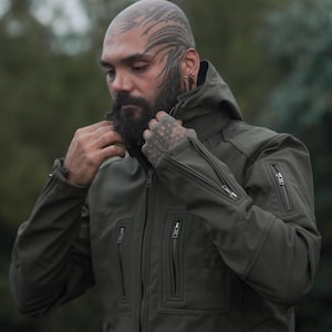 May include: An olive green hooded jacket with multiple zipped pockets. The jacket features a high collar and a fitted design. The model in the image is wearing the jacket. A versatile outerwear garment.