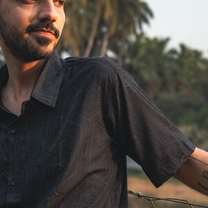 May include: A man wearing a black short-sleeved shirt with a geometric pattern. He is standing in front of a chain-link fence with trees in the background.