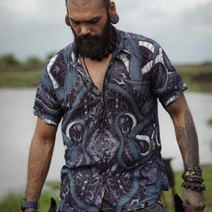 May include: A man with a beard and tattoos is wearing a blue and black patterned shirt with a button-down collar. He is holding two black dogs on leashes.